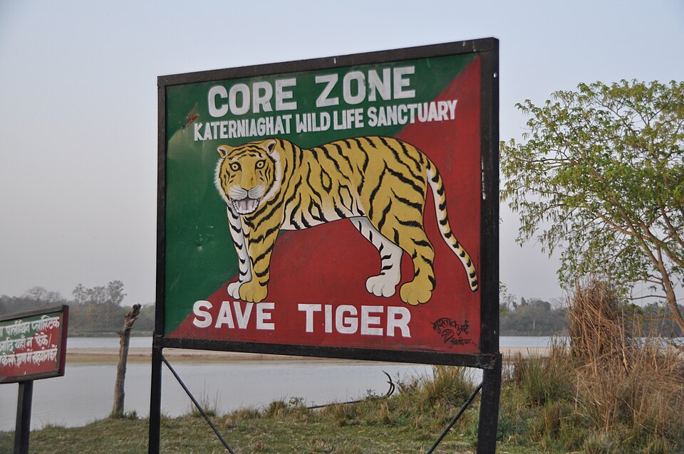 A painted signboard reading CORE ZONE, KATERNIAGHAT WILD LIFE SANCTUARY beside a broad river with grassland and forest on the far bank