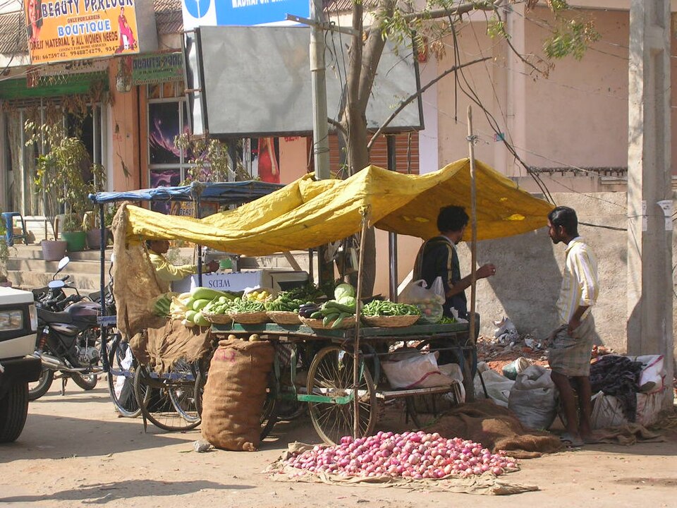 A street-side vegetable stall built on a handcart under a yellow tarpaulin, with baskets of gourds and beans on the cart and a heap of onions spread on the ground