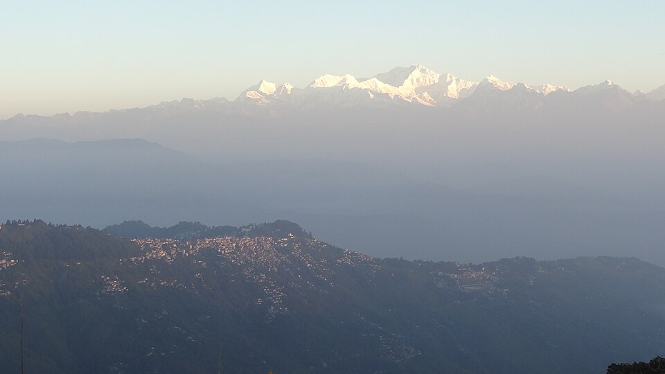 Darjeeling town spread along a forested ridge, with layered blue hills below and the snow-covered Kanchenjunga range on the skyline