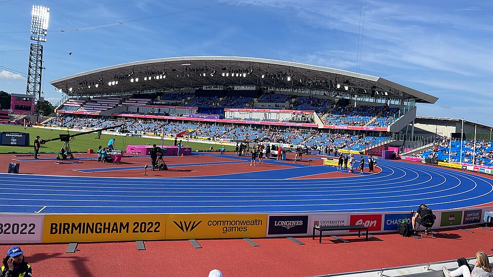 The blue running track and main grandstand of Alexander Stadium, with trackside hoardings reading Birmingham 2022 Commonwealth Games