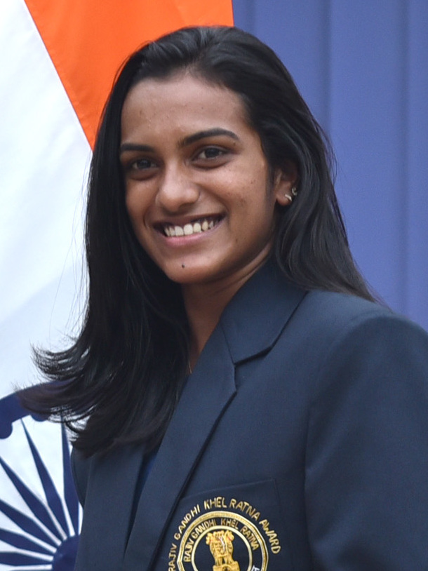 Head-and-shoulders portrait of the badminton player P. V. Sindhu smiling in a dark blazer bearing a Rajiv Gandhi Khel Ratna Award crest, the Indian tricolour behind her