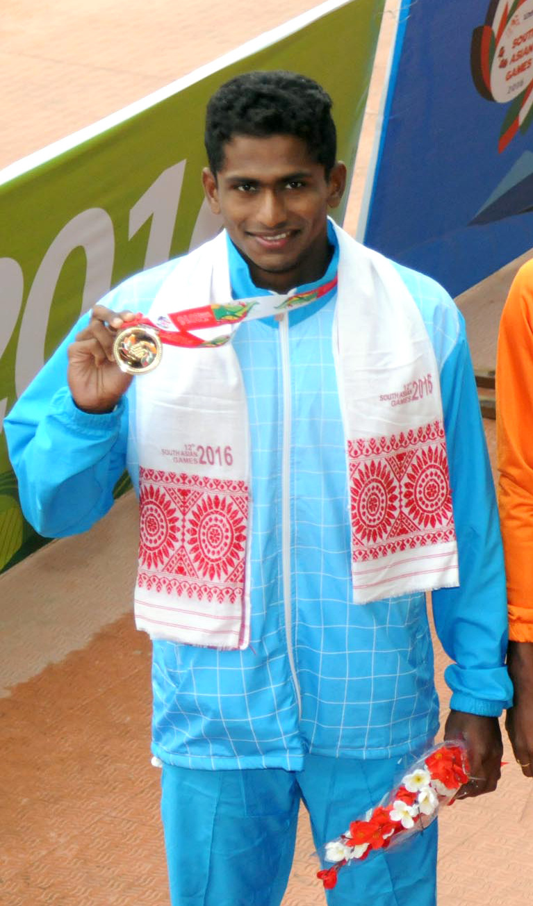 An Indian swimmer in a light-blue national tracksuit holding up a gold medal on a red ribbon at a medal ceremony