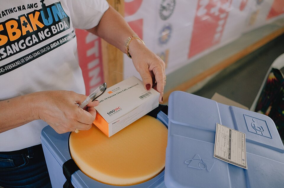 A health worker holding a Sinovac CoronaVac carton labelled COVID-19 Vaccine (Vero Cell), Inactivated, above a blue vaccine cold box