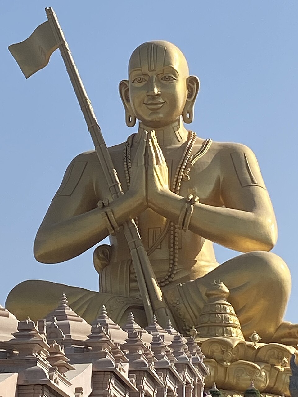 The golden seated Statue of Equality depicting Ramanuja with palms joined and a tridandi staff, rising above the spires of the temple platform below