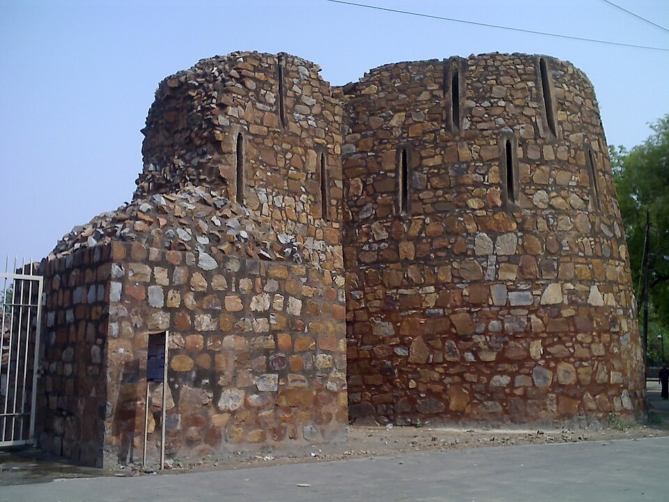 A ruined rubble-stone bastion of the Feroz Shah Kotla fort wall in Delhi, pierced by narrow arrow slits, standing beside a road