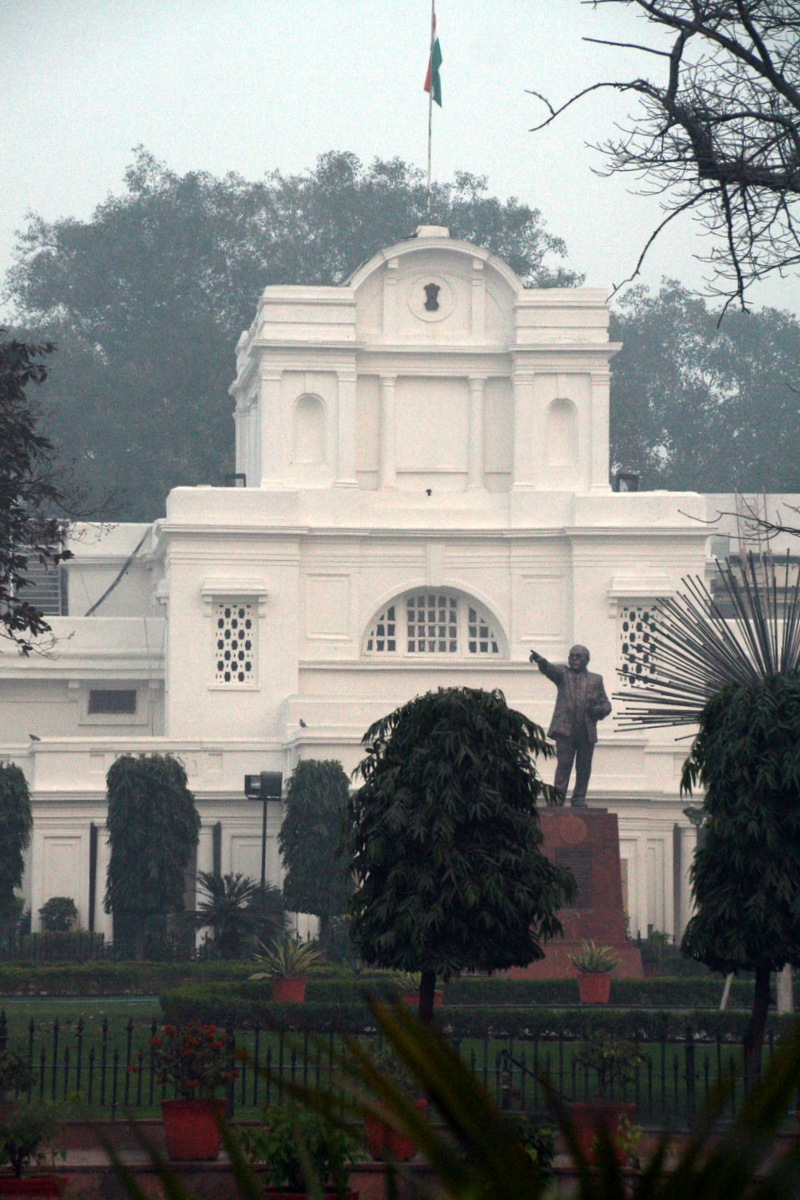 The white colonial-era Delhi Vidhan Sabha building behind garden hedges, the national flag on its roof and a statue of B. R. Ambedkar in the foreground