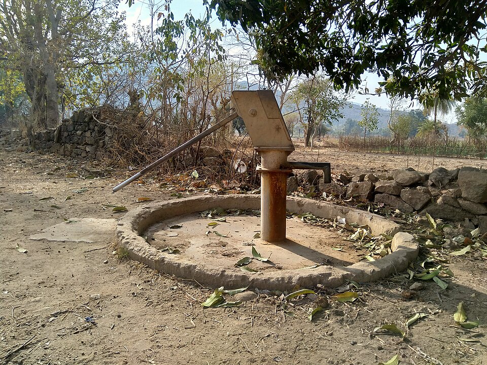 A cast-iron hand pump standing on a circular concrete apron over a shallow tube well beside a dry village track