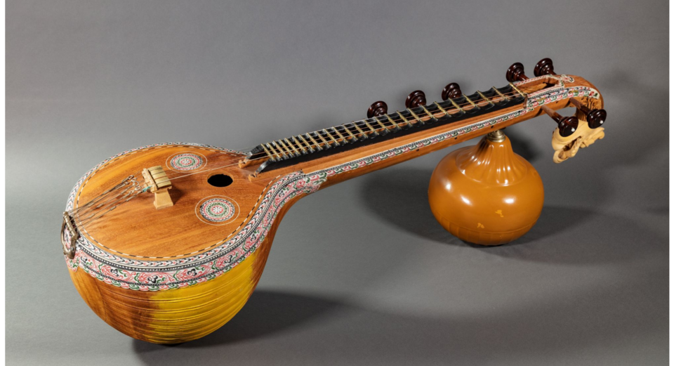 A Saraswati veena laid on its side: large decorated wooden resonator, long fretted neck with tuning pegs, and a second gourd under the carved yali head