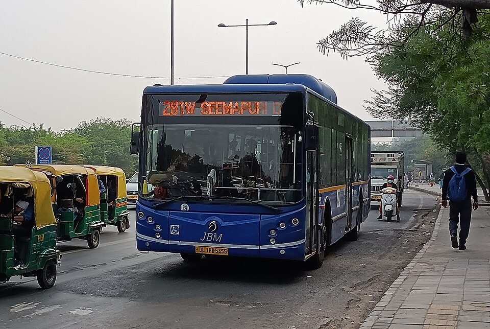 A blue CNG-powered low-floor city bus on route 281W travelling along an Indian arterial road beside auto-rickshaws