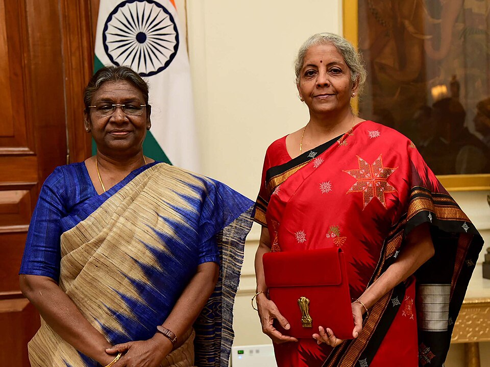 Finance Minister Nirmala Sitharaman in a red saree holding the red budget folder bearing the national emblem, standing beside President Droupadi Murmu in front of the Indian tricolour