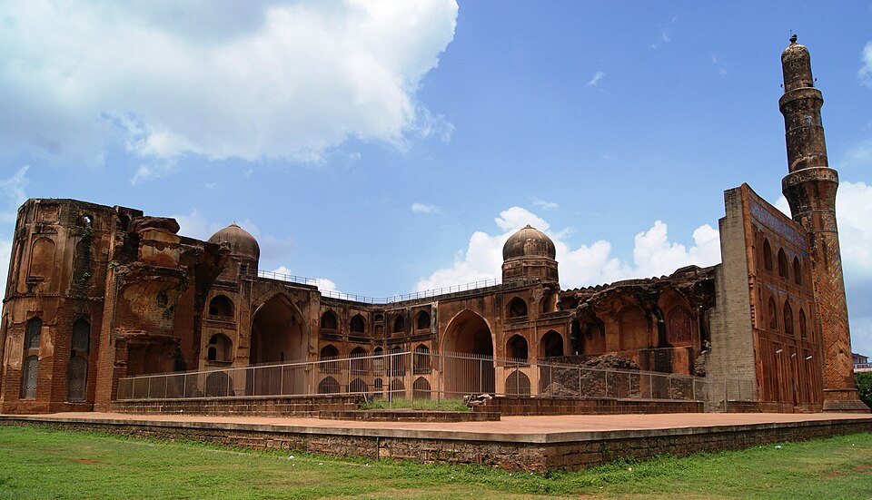 The ruined multi-storeyed Madrasa of Mahmud Gawan at Bidar, with rows of arched cells, domes and a surviving tall minaret against a blue sky
