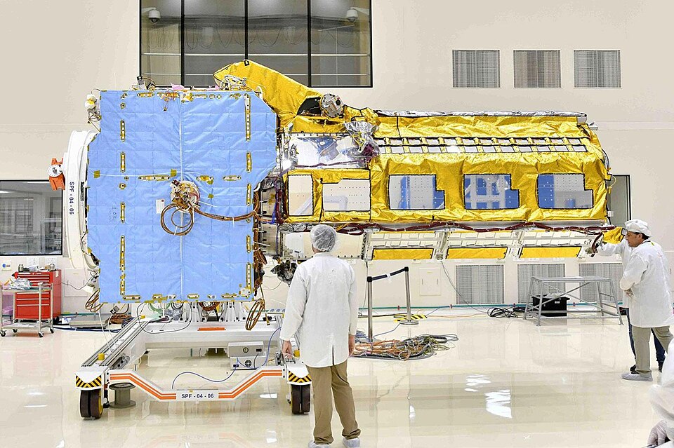 The NISAR (NASA-ISRO Synthetic Aperture Radar) satellite under assembly in a clean room, its gold-foil-wrapped body and folded blue solar panel flanked by engineers in white coats