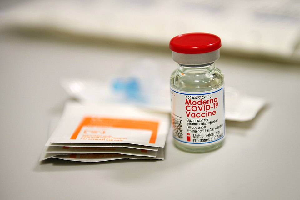 A single glass vial with a red cap labelled Moderna COVID-19 Vaccine standing beside alcohol prep pads on a white surface