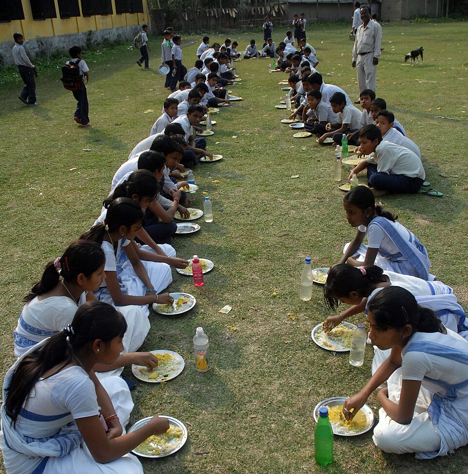 Two long rows of uniformed schoolchildren seated on the ground of a school yard eating a cooked mid-day meal from plates