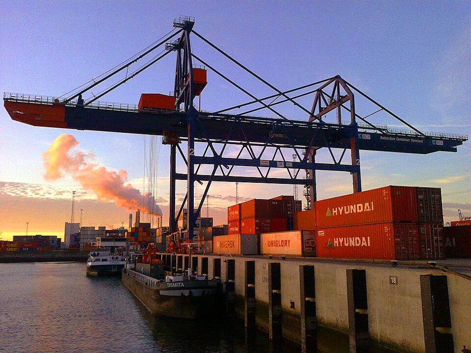 A gantry crane marked Rotterdam Container Terminal towering over stacked shipping containers and a barge at the Port of Rotterdam at sunset