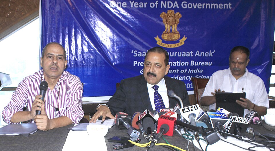 Dr Jitendra Singh (centre) in a dark suit and tie speaking at a Press Information Bureau press conference table lined with news microphones, flanked by two other men