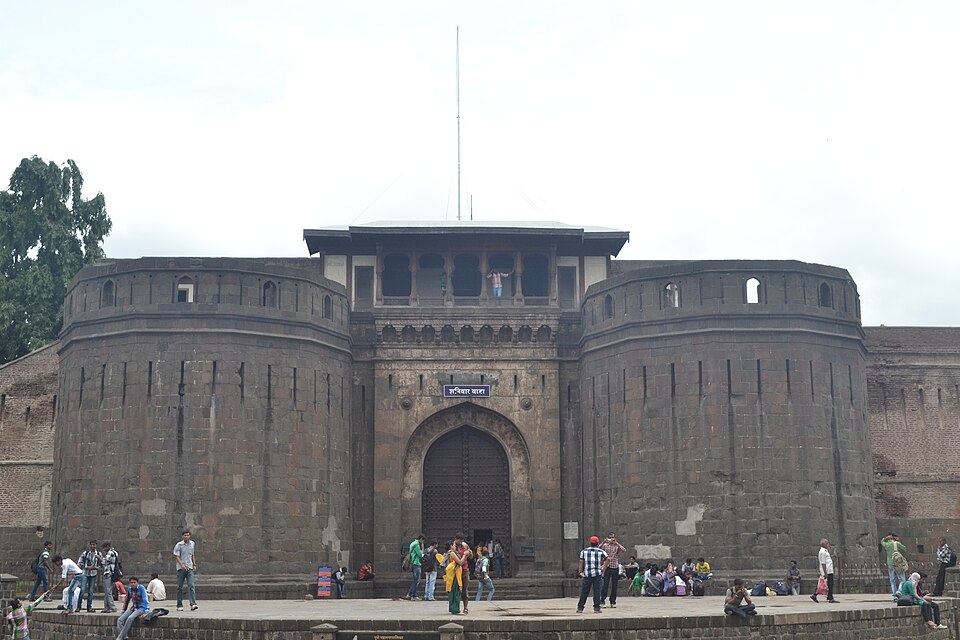 The dark stone main gateway of Shaniwar Wada in Pune, flanked by two rounded bastions, with a studded wooden door set in a pointed arch and visitors on the steps in front