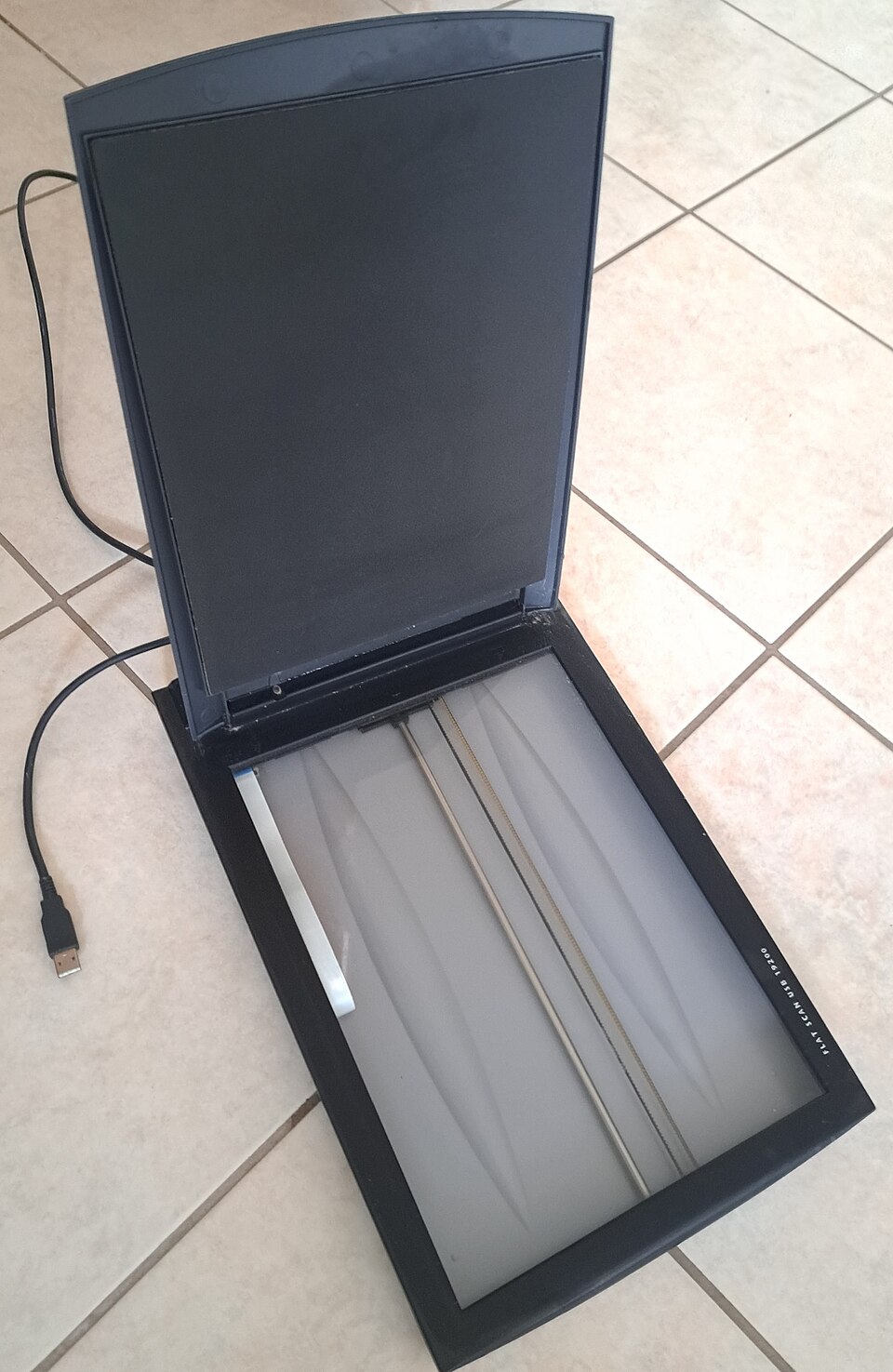 A black flatbed document scanner lying open on a tiled floor with its hinged lid raised, showing the rectangular glass platen and the scanning head bar underneath it, and a USB cable running from the back with the unplugged connector in the foreground