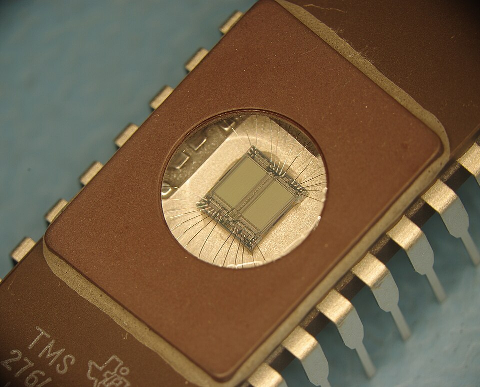 A close macro photograph of an EPROM integrated circuit: a rectangular ceramic chip package with two rows of flat metal legs along its sides and a round transparent quartz window set into the top, through which the silvery silicon die and its fine bond wires can be seen