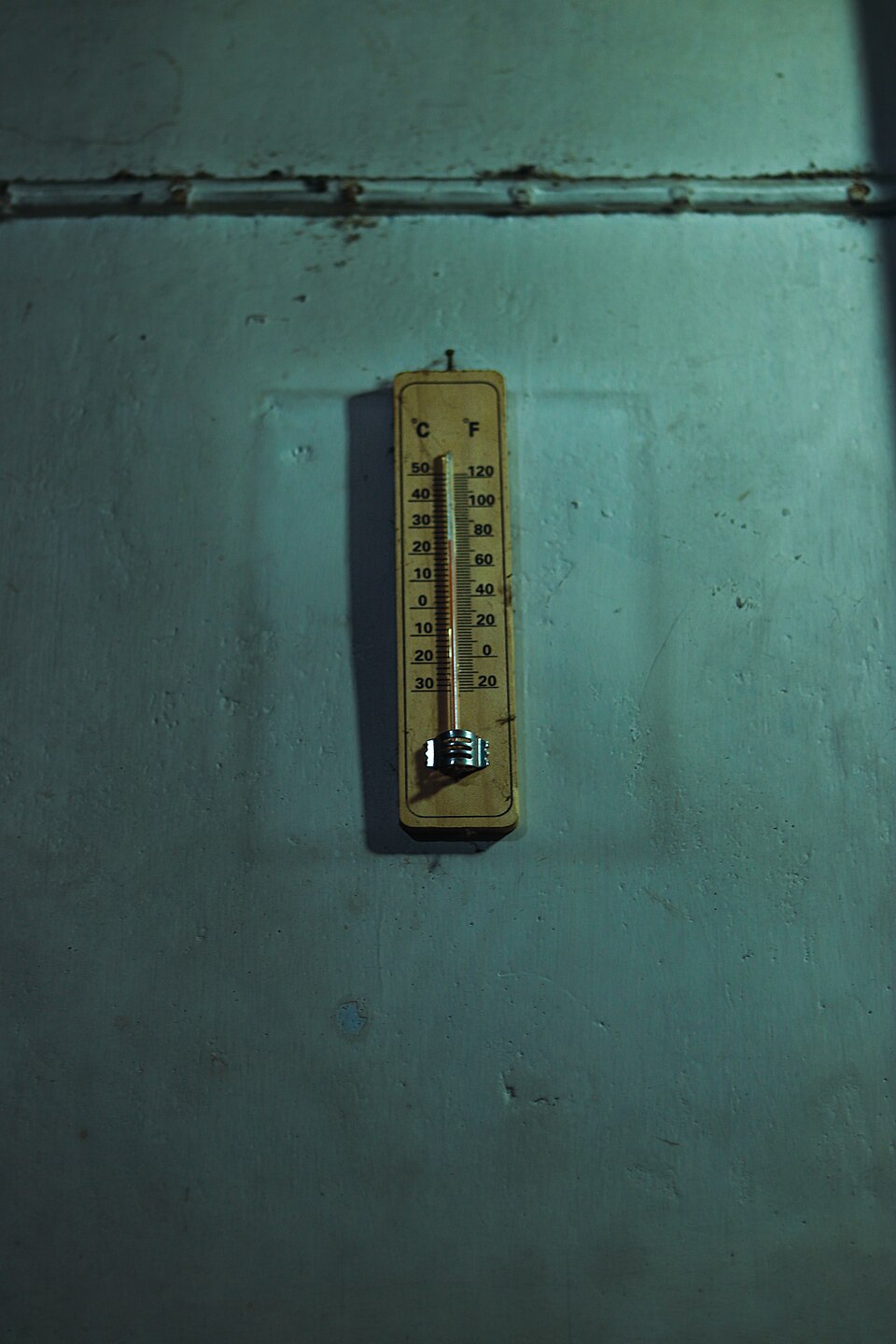 A small wooden-backed wall thermometer hanging on a dim green-painted wall, its glass capillary flanked by two printed scales — degrees Celsius numbered 50 down to minus 30 on the left and degrees Fahrenheit numbered 120 down to minus 20 on the right — so the two readings line up against each other
