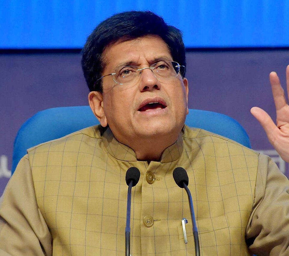 Piyush Goyal, a bespectacled man in a beige checked Nehru jacket, speaking into two table microphones with one hand raised at a press conference