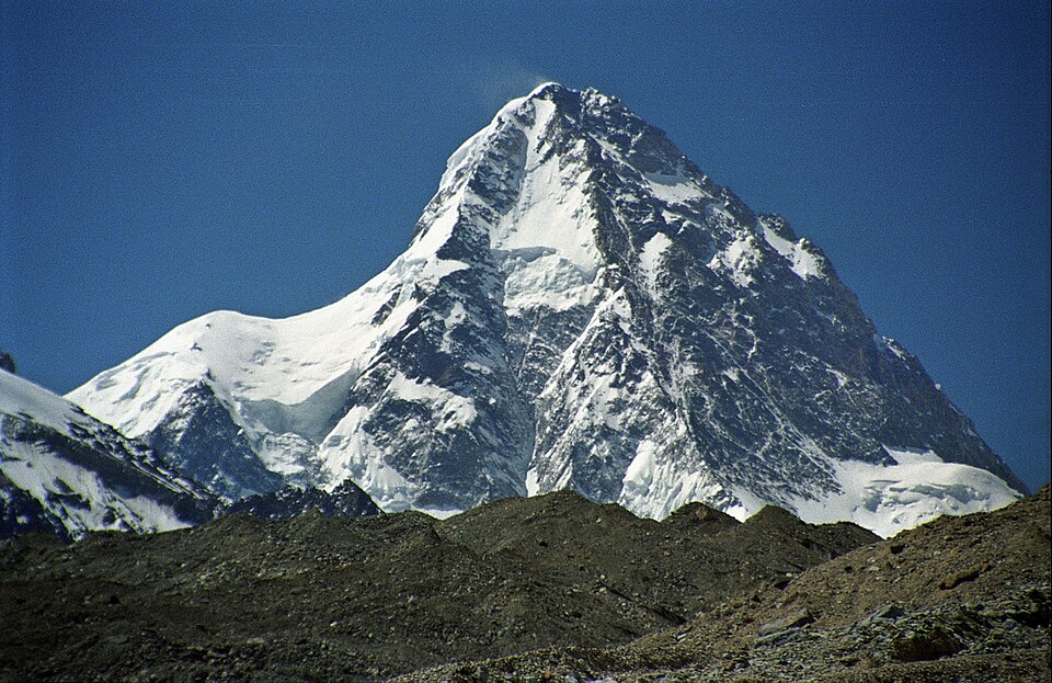 A steep pyramid-shaped snow and rock peak rising against a deep blue sky above a dark grey glacial moraine ridge, with hanging glaciers on its flanks