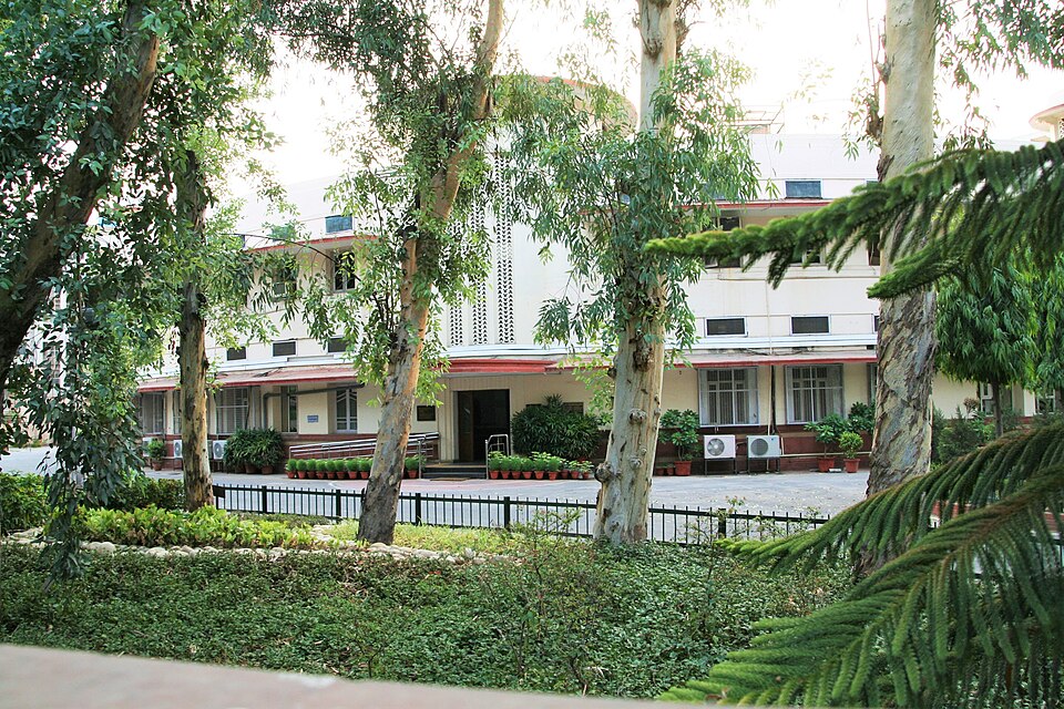 A long white two-storey Art Deco building with red-brown eaves, a covered porch entrance and rows of potted plants along its front, seen from a garden through the trunks of tall eucalyptus trees