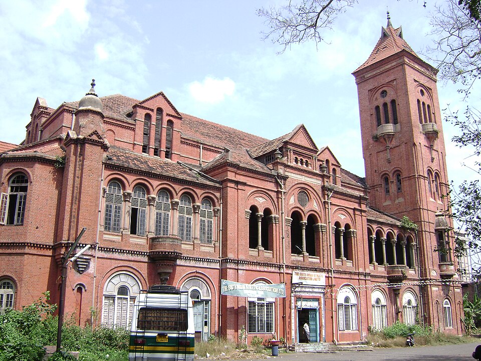 The red-brick Victoria Public Hall in Chennai, a two-storey Indo-Saracenic building with arcaded verandas, gabled tiled roofs and a tall corner tower with a pointed spire, seen from the street