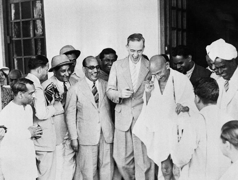 A black-and-white 1942 photograph of Sir Stafford Cripps in a light suit standing on a doorstep laughing beside Mahatma Gandhi, who is wrapped in a white khadi shawl, with a crowd of smiling Indian and British men around them