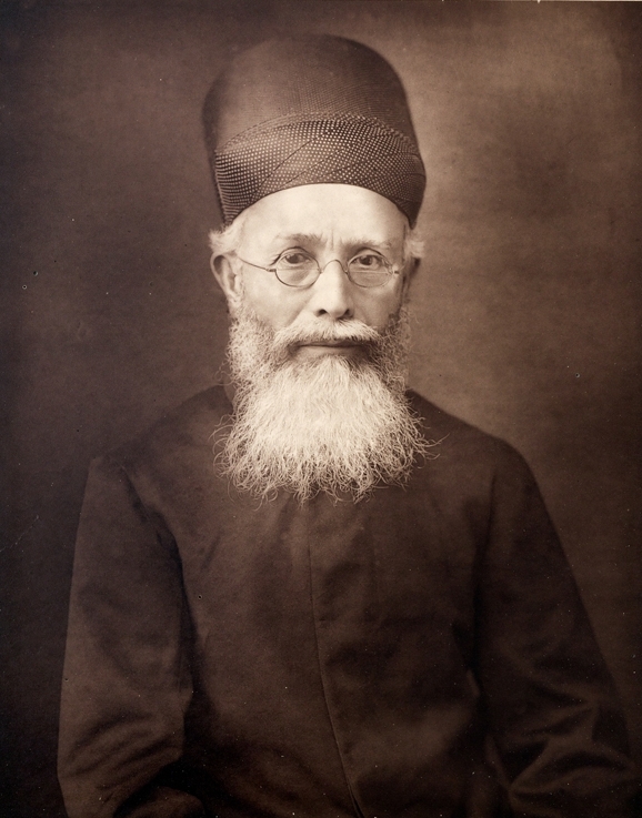 A sepia studio portrait of Dadabhai Naoroji as an elderly man in a tall Parsi cap, round wire spectacles and a long white beard, wearing a dark high-collared coat