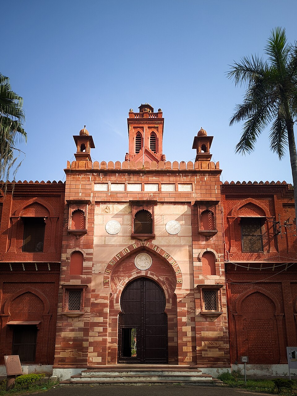 A red sandstone and brick gateway with a tall pointed archway, the words VICTORIA GATE carved in a curve above the black wooden doors, flanked by domed turrets and a small clock tower behind, with palm trees on either side against a blue sky
