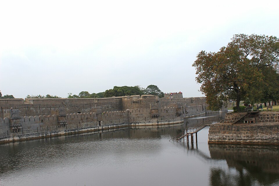 A long low rampart of grey granite blocks running along the far bank of a wide still moat, with trees growing behind the wall and a small footbridge crossing the water on the right