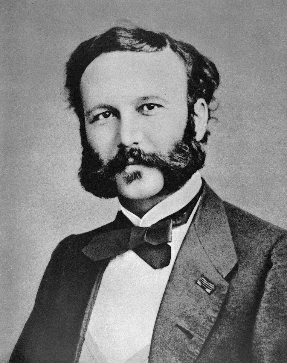 A nineteenth-century black-and-white studio portrait of Henry Dunant as a young man with dark wavy hair, a full beard and moustache, wearing a large bow tie, waistcoat and jacket with a small ribbon badge on the lapel