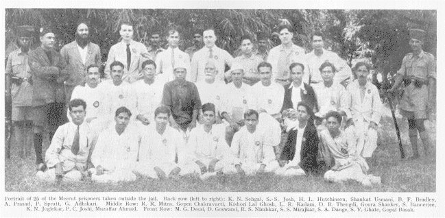 A black-and-white group photograph of twenty-five men, several rows standing and one row seated on the ground, flanked by uniformed guards outside a jail, under a printed caption naming them as the Meerut prisoners