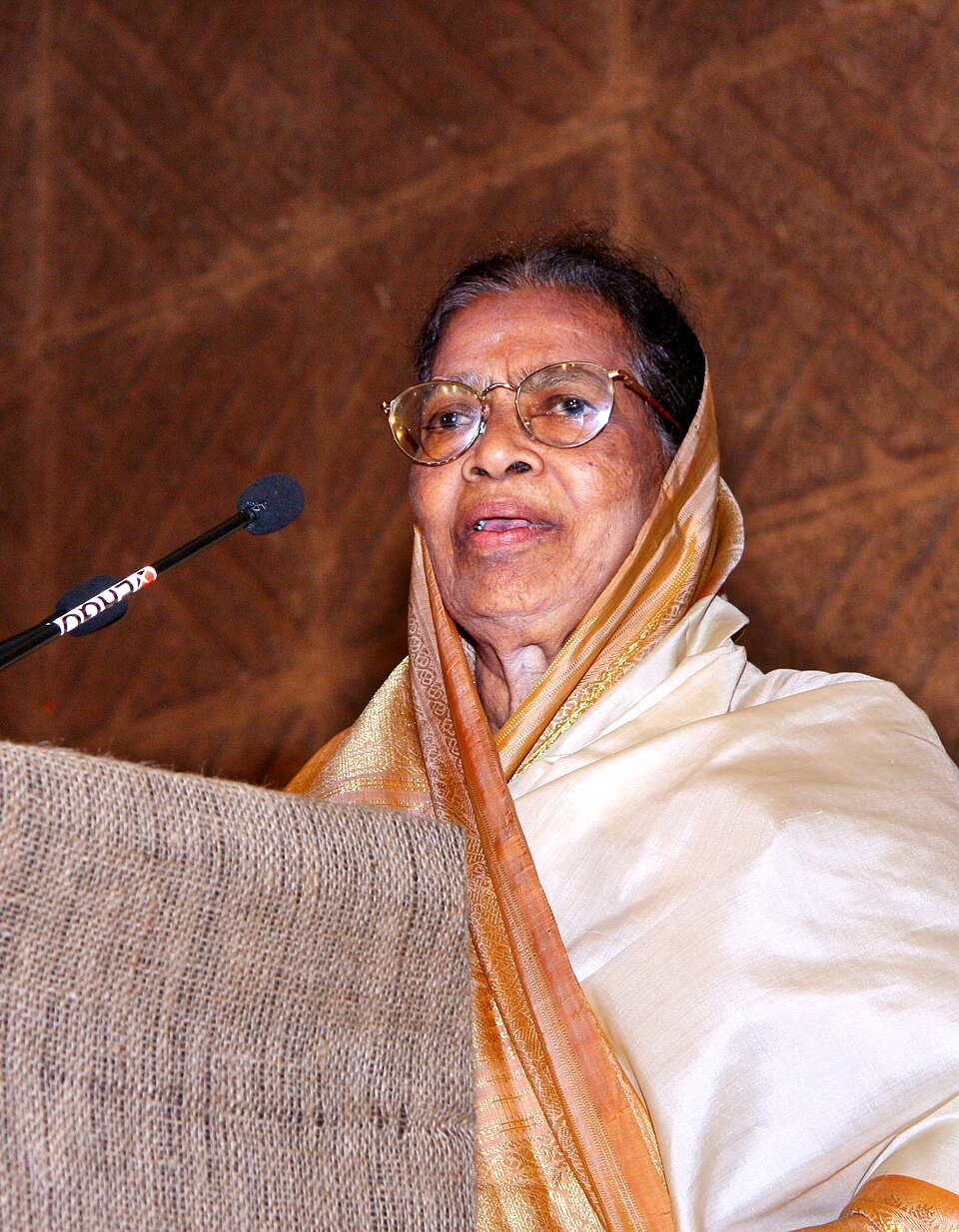 An elderly woman in a cream and gold-bordered sari with the end drawn over her head, wearing gold-rimmed spectacles, speaking from behind a jute-covered lectern into a microphone