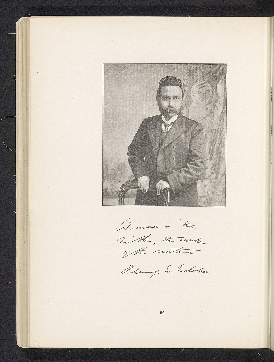 An album page carrying a mounted studio photograph of Behramji Malabari, a moustached man in a Parsi cap and dark frock coat standing behind a chair, above a handwritten inscription signed with his name