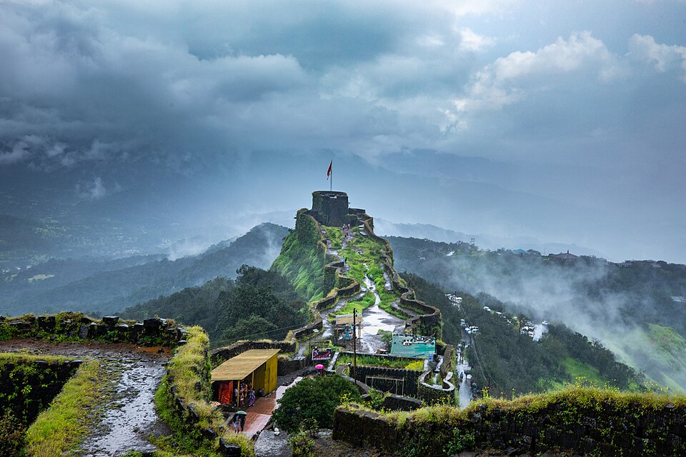 Pratapgad fort in the monsoon, its stone ramparts stepping up a green ridge to an upper citadel flying a saffron flag, with mist and the Sahyadri hills behind