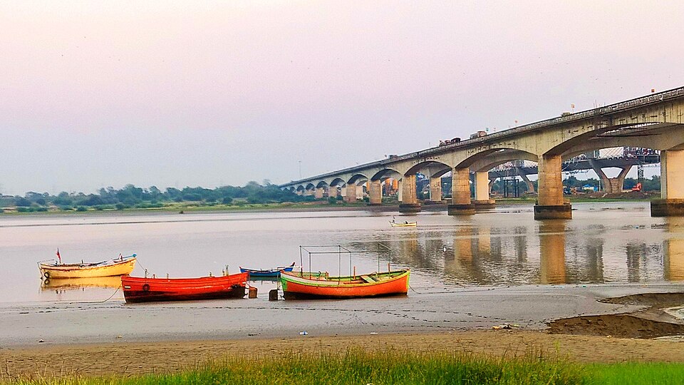Wooden fishing boats drawn up on the mud bank of the broad Narmada river at Bharuch at dusk, with a long arched road bridge crossing the water behind them