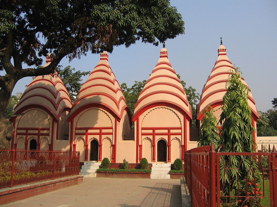 A row of four matching cream-and-red Bengali-style temples with ridged conical spires and arched doorways, standing behind a red railing in the Dhakeshwari temple compound at Dhaka