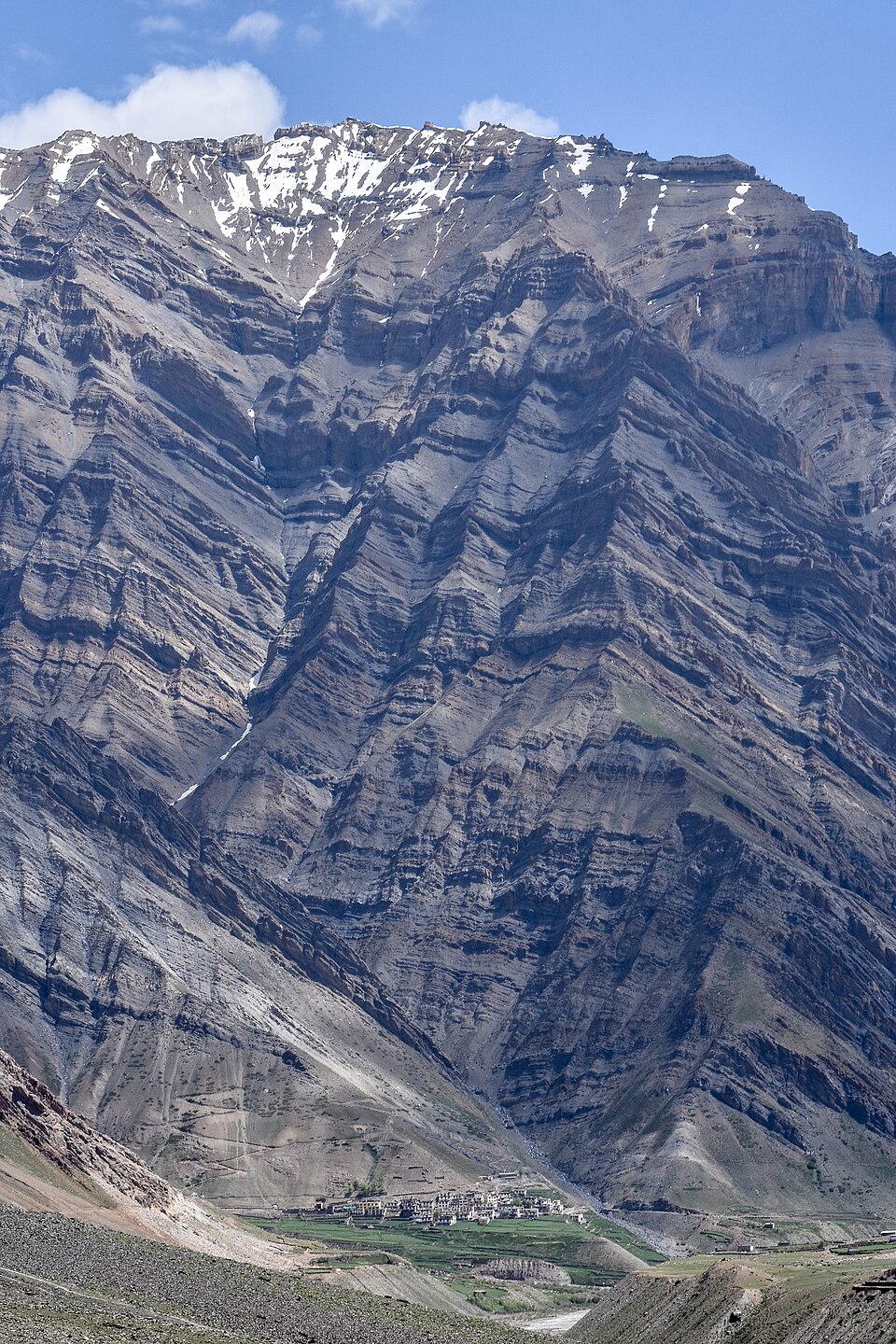 A vast bare, layered rock mountainside streaked with snow rising above a small stone village and a strip of green fields on the valley floor in the Pin valley of Spiti, with no trees anywhere on the slopes