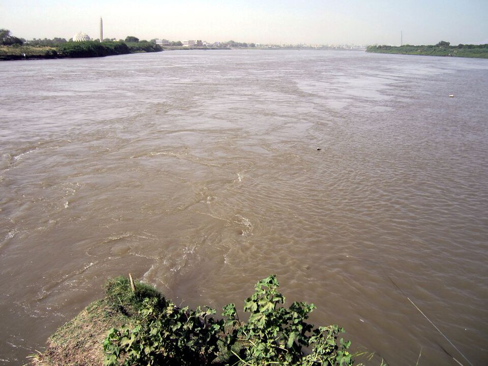 Two broad channels of muddy brown river water swirling together where the Blue and White Nile meet at Khartoum, seen from a low vegetated bank, with the citys buildings and a tall tower on the far shore