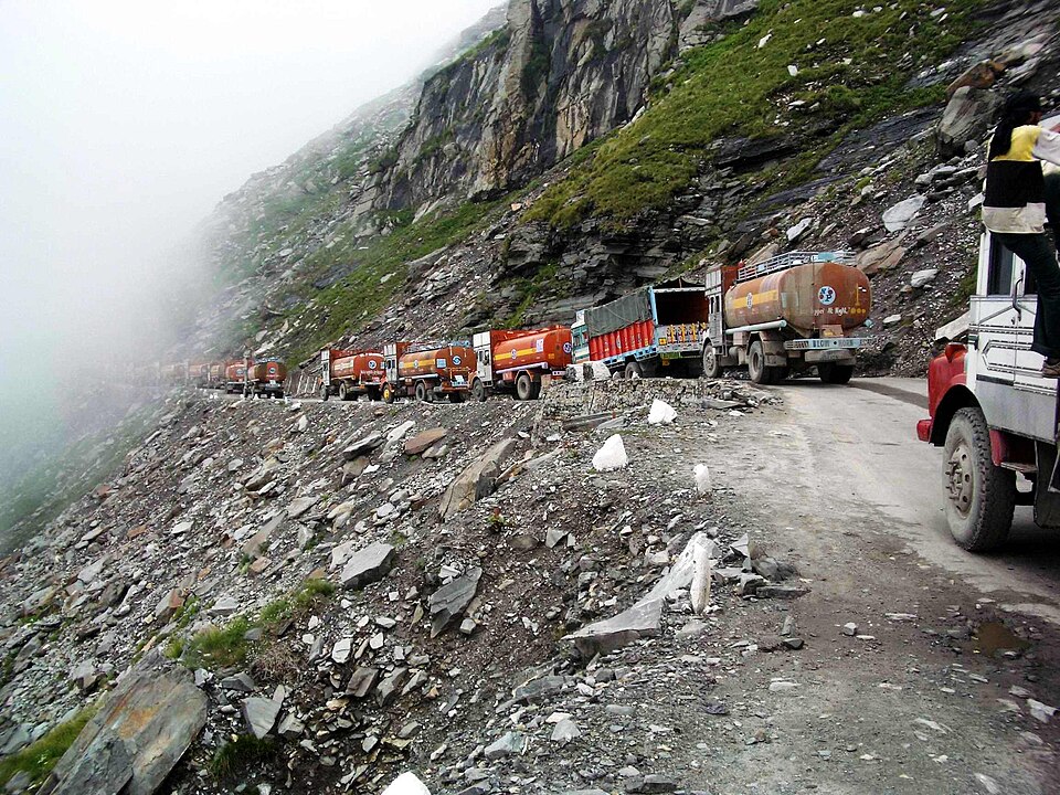 A line of tanker trucks queued along a narrow unpaved road cut into a misty, scree-covered mountain slope on the Rohtang Pass crossing in Himachal Pradesh