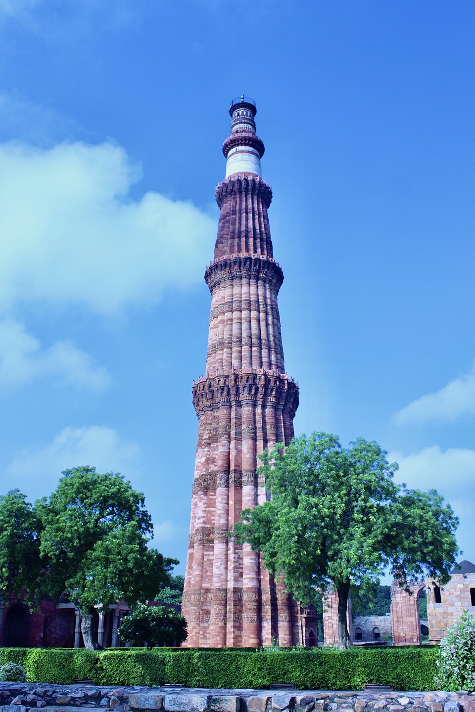 The Qutb Minar in Delhi seen from its base against a blue sky - a tapering red sandstone tower with fluted, balconied storeys and a pale marble upper section, rising above trees and the ruined arcades of the surrounding complex