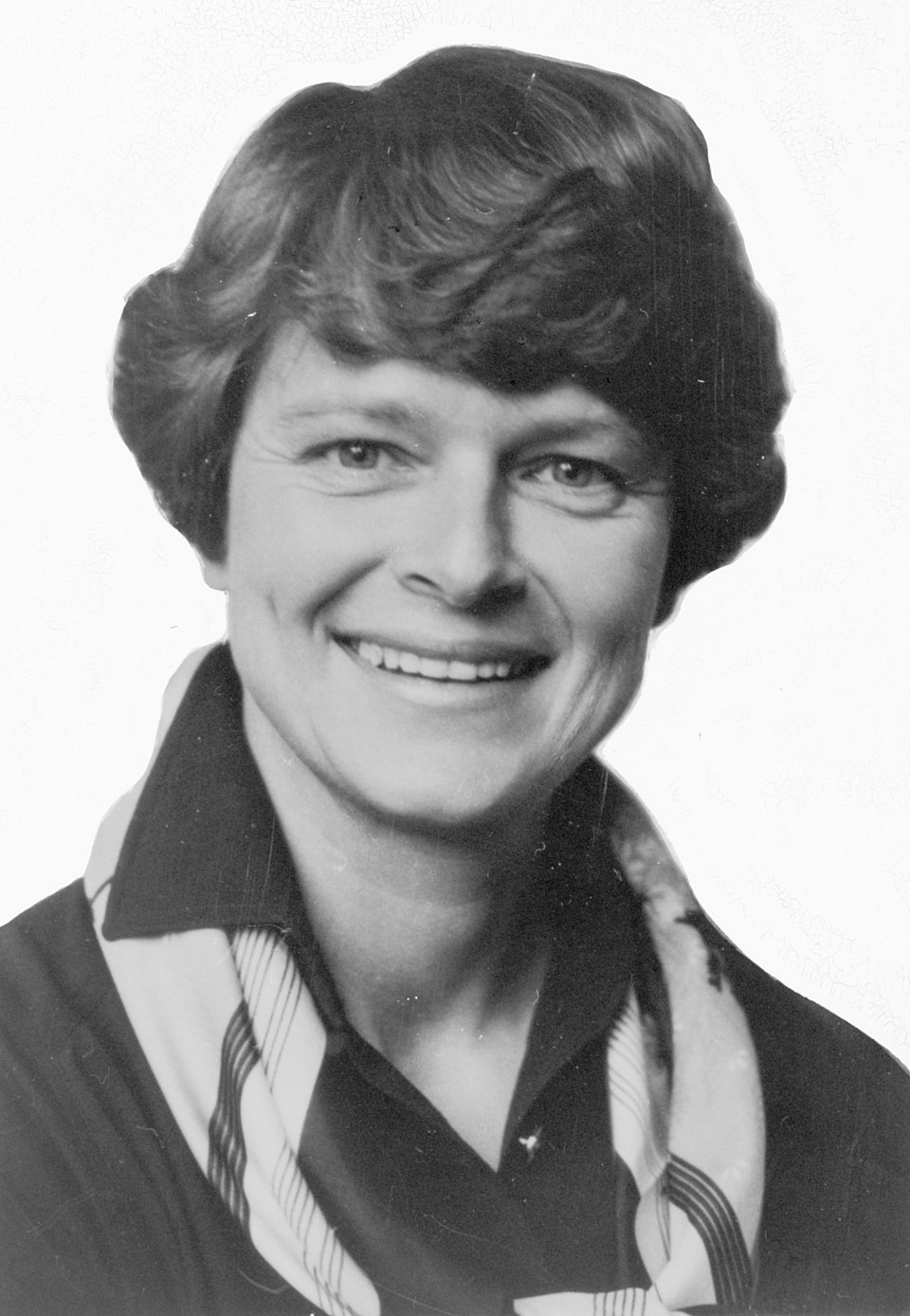 A black-and-white studio portrait of Gro Harlem Brundtland in the 1970s, a smiling woman with short wavy hair wearing a dark jacket over a patterned scarf