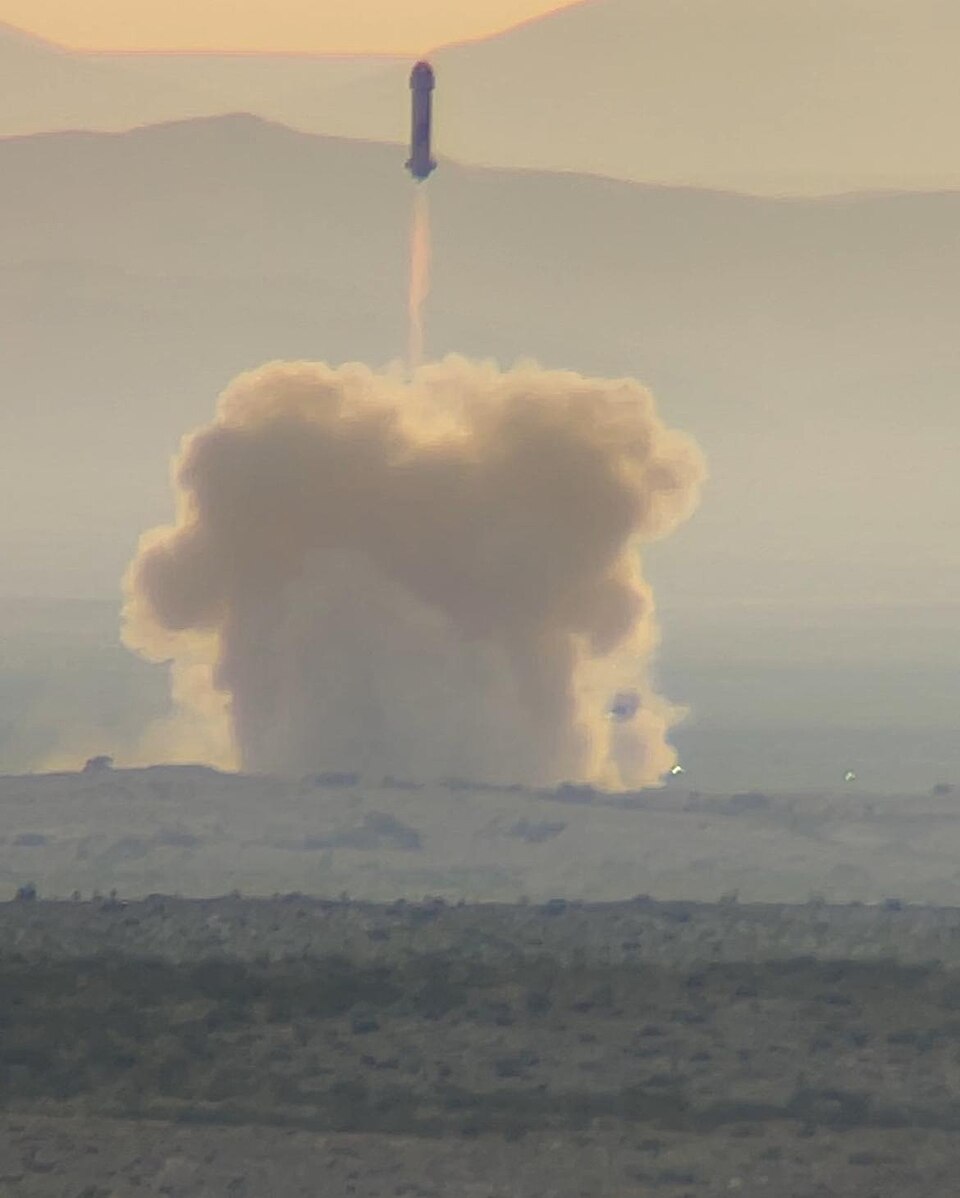 A Blue Origin New Shepard rocket climbing on a bright exhaust plume above the West Texas desert, with a large pale dust cloud billowing across the launch site and low hills in the haze behind