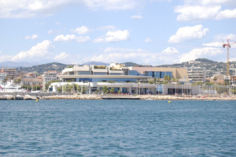 The Palais des Festivals et des Congres at Cannes seen from the sea, a low white and glass building with a curved roof canopy on the waterfront, with the town, moored yachts and wooded hills behind it