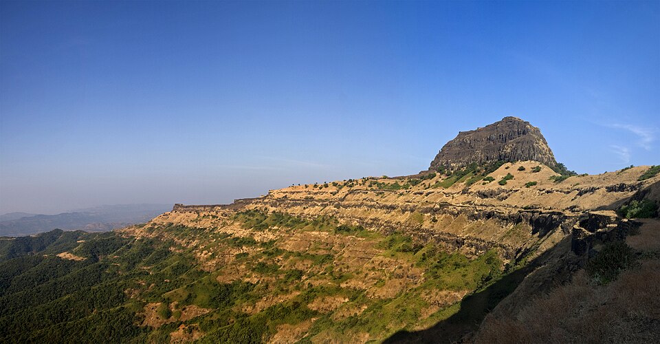 A flat-topped dark basalt hill above a long ridge in the Maharashtra Ghats, with horizontal rock ledges stepping one above another across the grassy slopes and a stone rampart following the crest