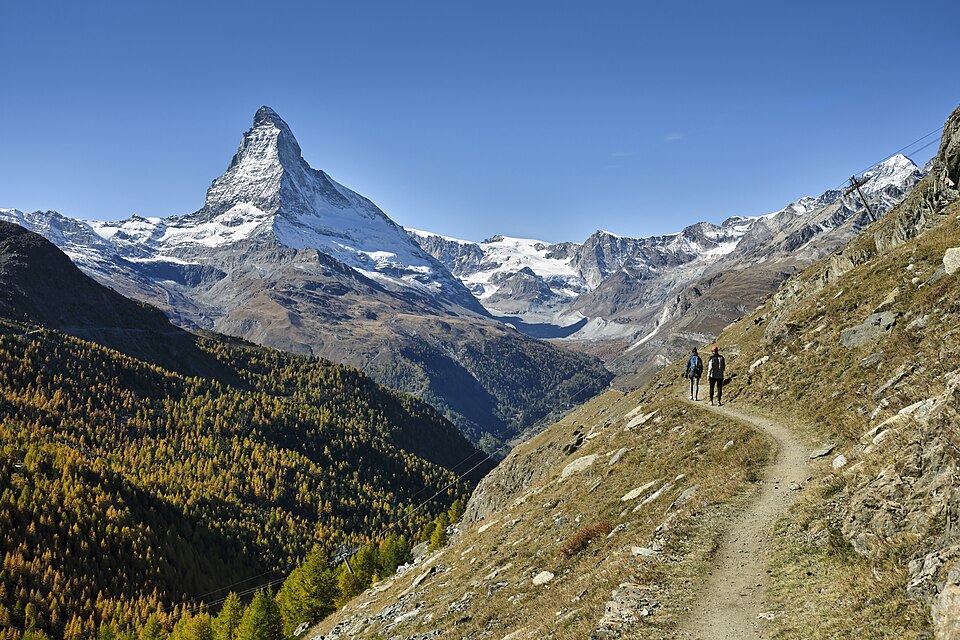 The sharp snow-streaked pyramid of the Matterhorn rising above a glaciated valley near Zermatt, with golden autumn larch forest on the left slope and two walkers on a path in the foreground