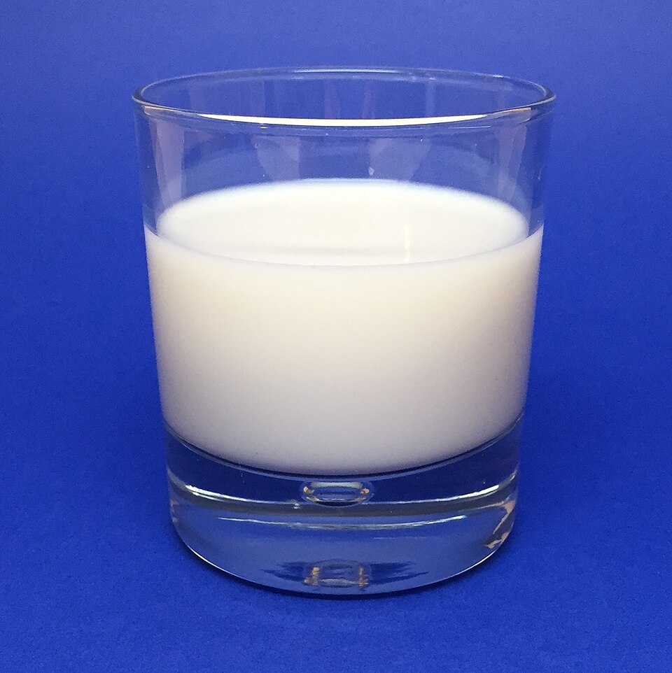 A clear drinking glass filled with opaque white milk, standing on a plain blue background