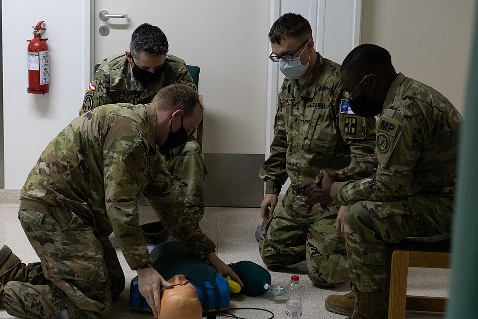 A kneeling trainee pressing down with both hands and straight arms on the chest of a CPR training manikin lying on the floor, while three others watch from beside it